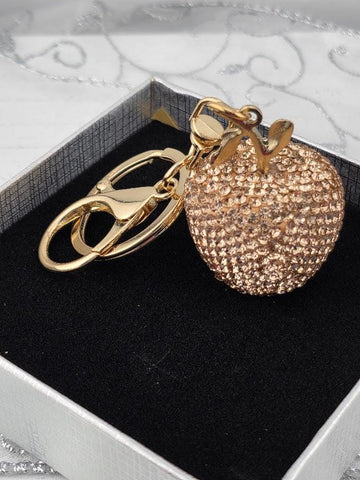 A gold-colored keychain in the shape of an apple, with a sparkling crystal-like design, is displayed in a black box.