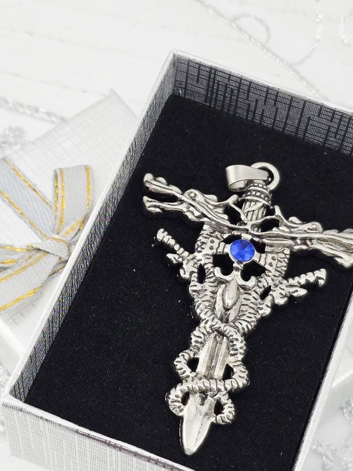 A silver-colored pendant with intricate designs and a blue gemstone is displayed in a black box with a gold ribbon.