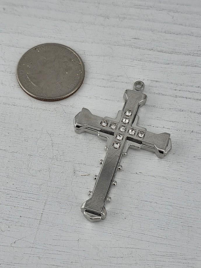 A silver cross pendant with small rhinestones is placed next to a silver coin on a white wooden surface.