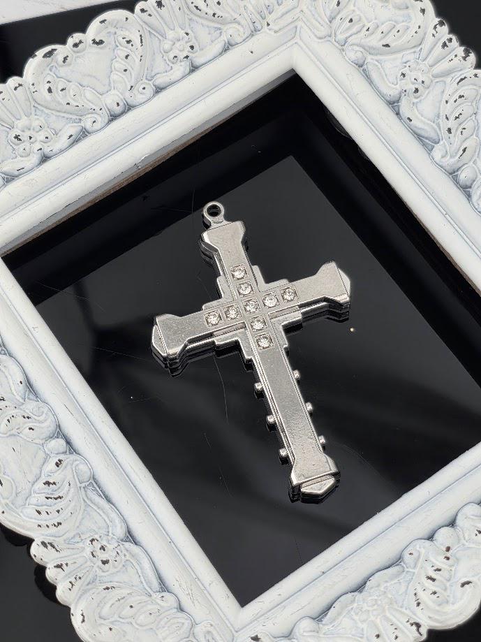 A silver cross-shaped pendant with small rhinestones or crystals is displayed in a white ornate frame against a black background.
