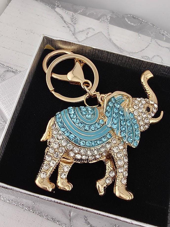 A decorative keychain in the shape of an elephant, with blue and gold accents, is displayed in a black box.