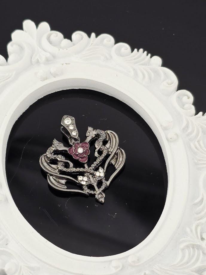 A decorative pendant with a floral design and a gemstone is displayed in a white ornate frame against a dark background.