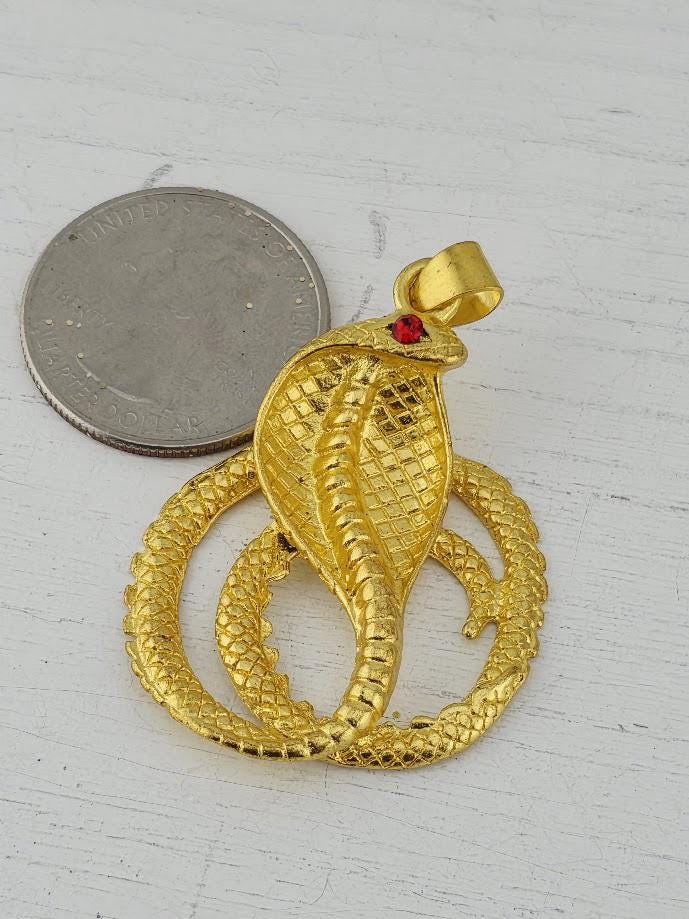 A gold pendant in the shape of a cobra, with a red gemstone in its eye, is placed next to a United States quarter coin on a white surface.