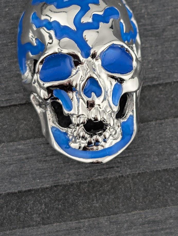 A silver and blue pendant in the shape of a skull is displayed in a white box, with a blue gift box and silver ribbon visible in the background.