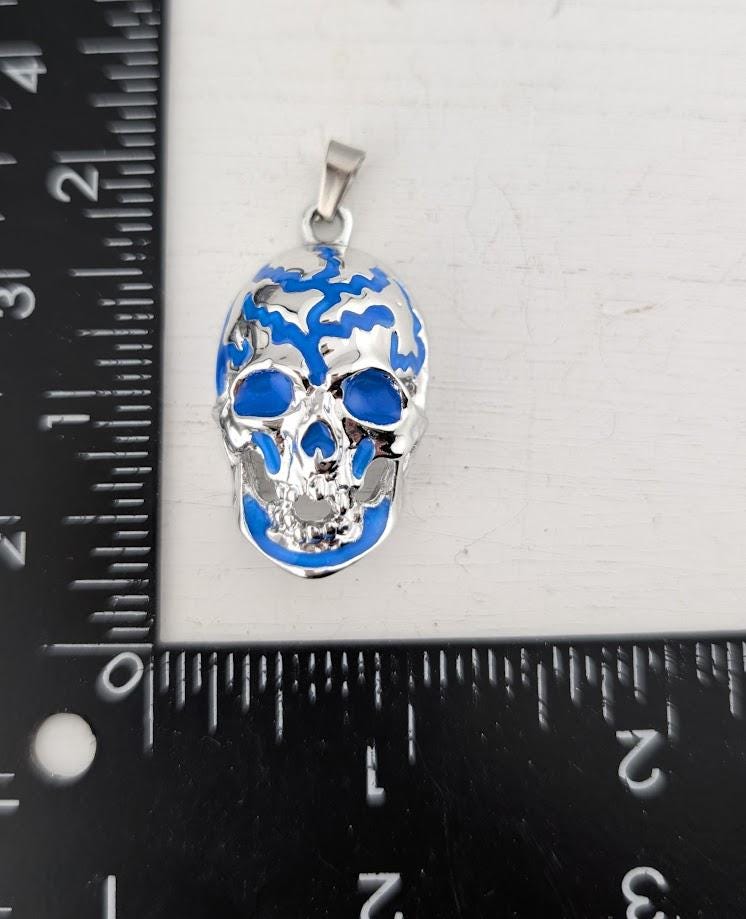 A blue and silver pendant in the shape of a skull is placed next to a ruler on a white surface.