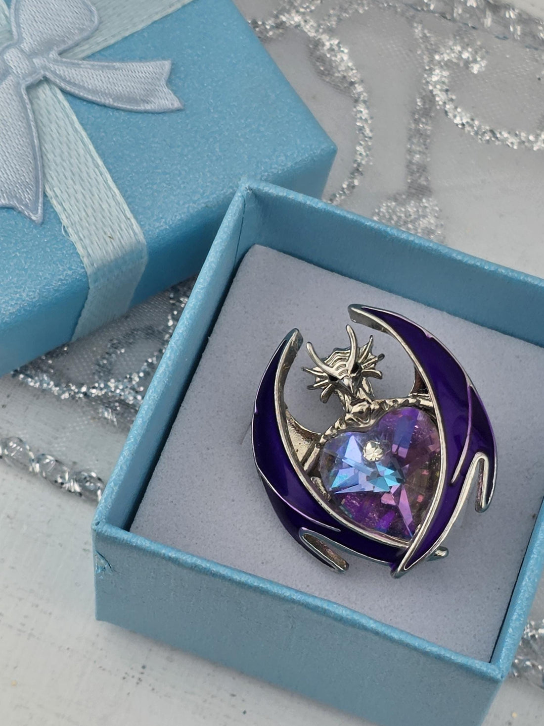 A purple brooch with a crescent moon design is displayed in a blue gift box with a white ribbon.