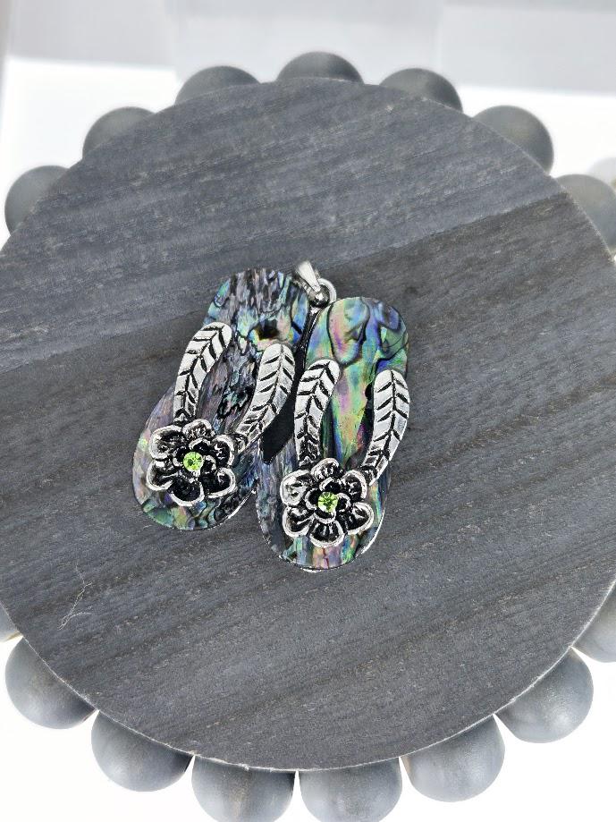 A pair of colorful, intricately designed flip-flop-shaped earrings with green stones, displayed in a pink box with a bow.