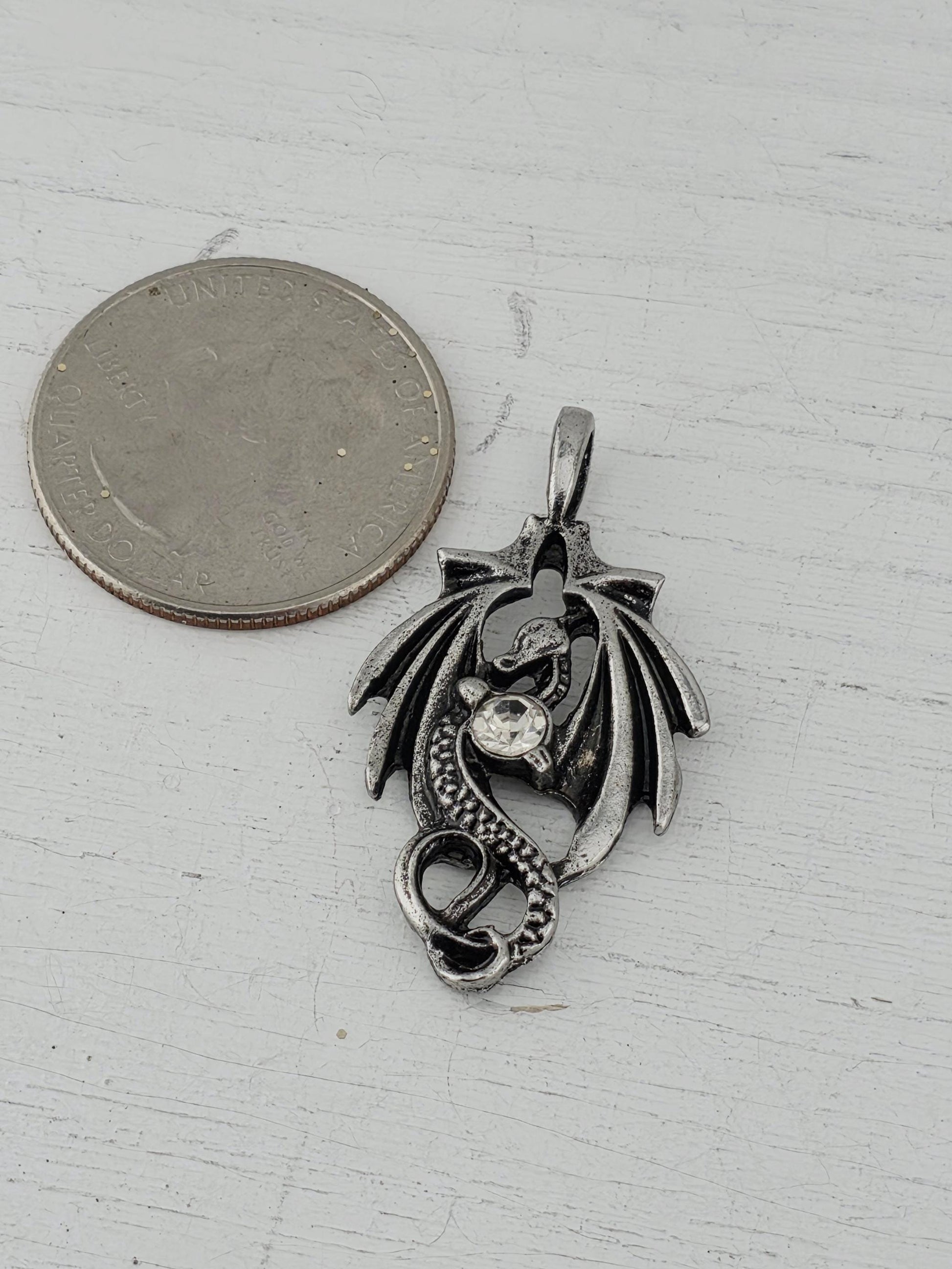 A silver-colored metal pendant in the shape of a dragon is placed next to a United States quarter on a white surface.