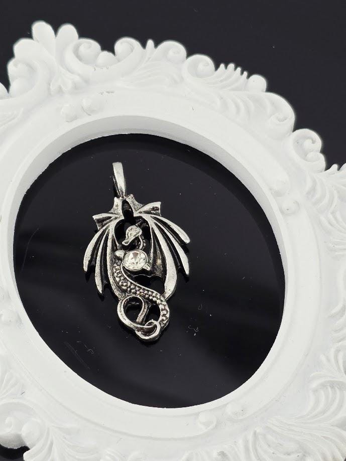 A silver pendant in the shape of a dragon is displayed against a black background, with a white ornate frame surrounding it.