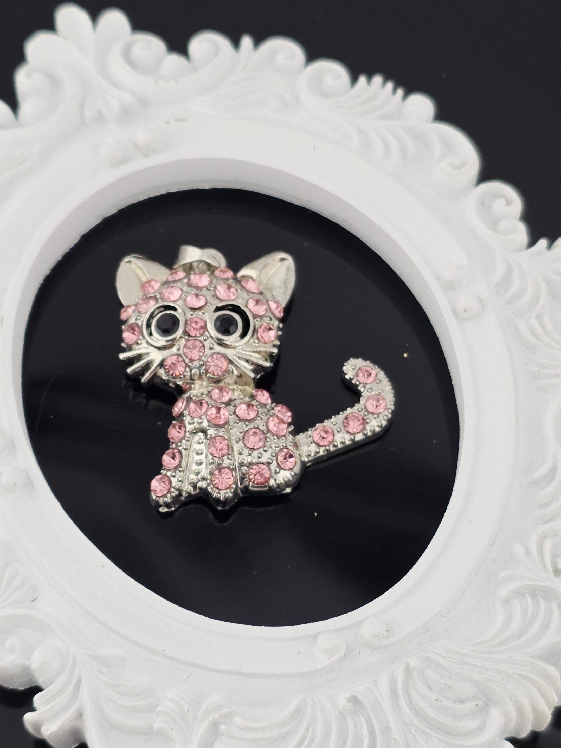 A decorative cat brooch with pink gemstones is displayed in a white ornate frame.