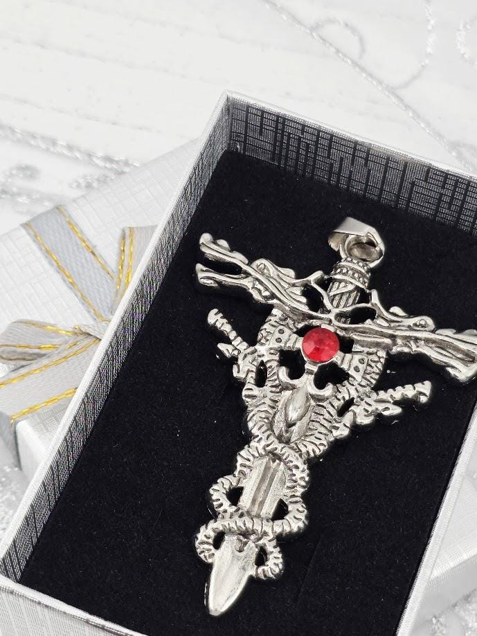A silver-colored pendant with a red gemstone, resembling a cross or sword, is displayed in a black box with a gold ribbon.
