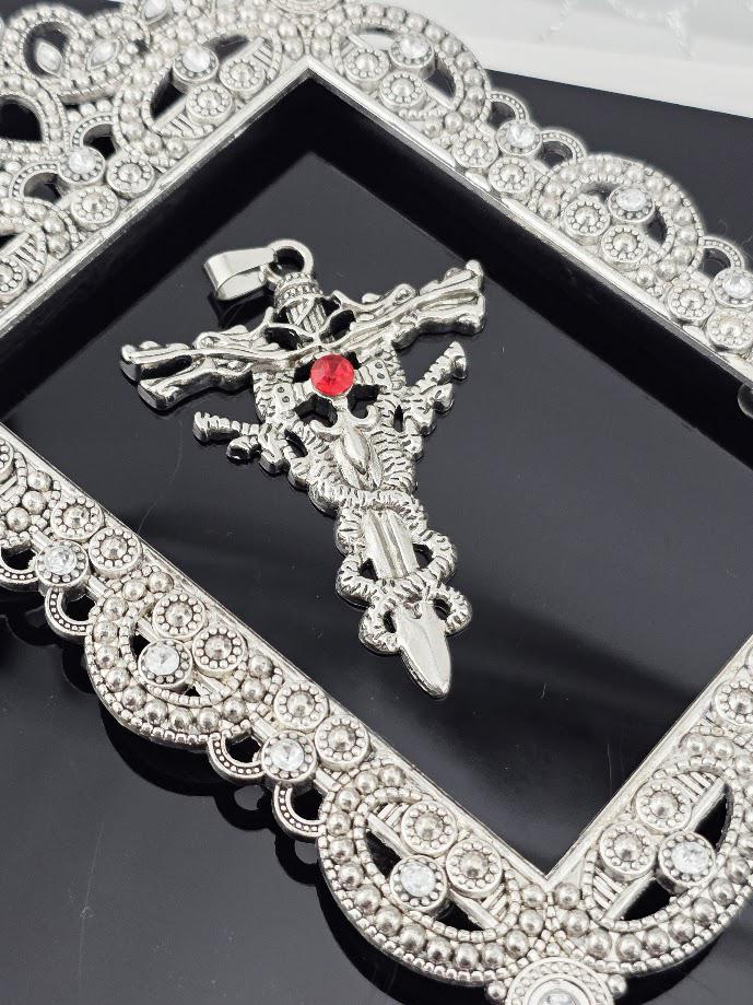 A silver-colored pendant with a red gemstone, resembling a cross or sword, is displayed in a black box with a gold ribbon.
