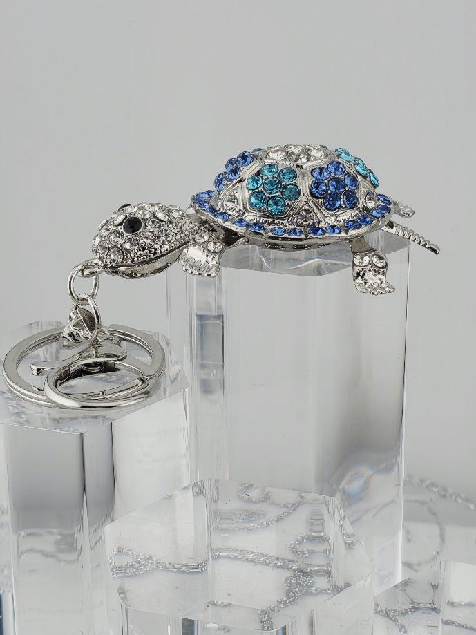 A silver keychain in the shape of a turtle, adorned with blue and white gemstones, is displayed on a clear glass stand.