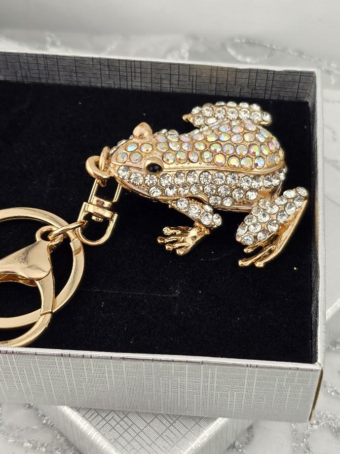 A gold-colored keychain in the shape of a frog, adorned with rhinestones or crystals, is displayed in a black box.