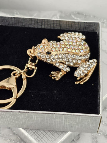 A gold-colored keychain in the shape of a frog, adorned with rhinestones or crystals, is displayed in a black box.