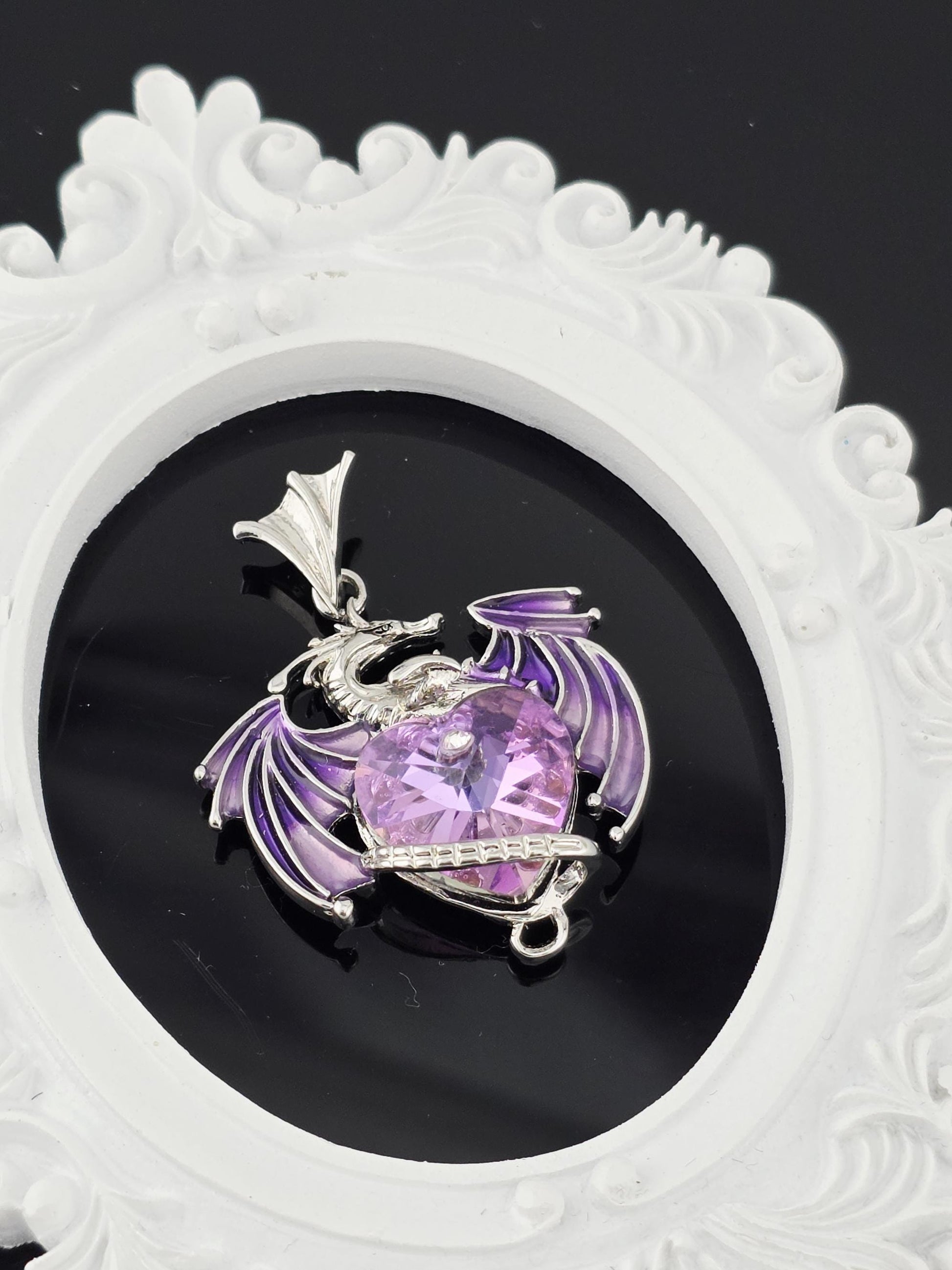 A piece of jewelry with a purple gemstone and a silver dragon design, displayed on a white ornate frame against a black background.