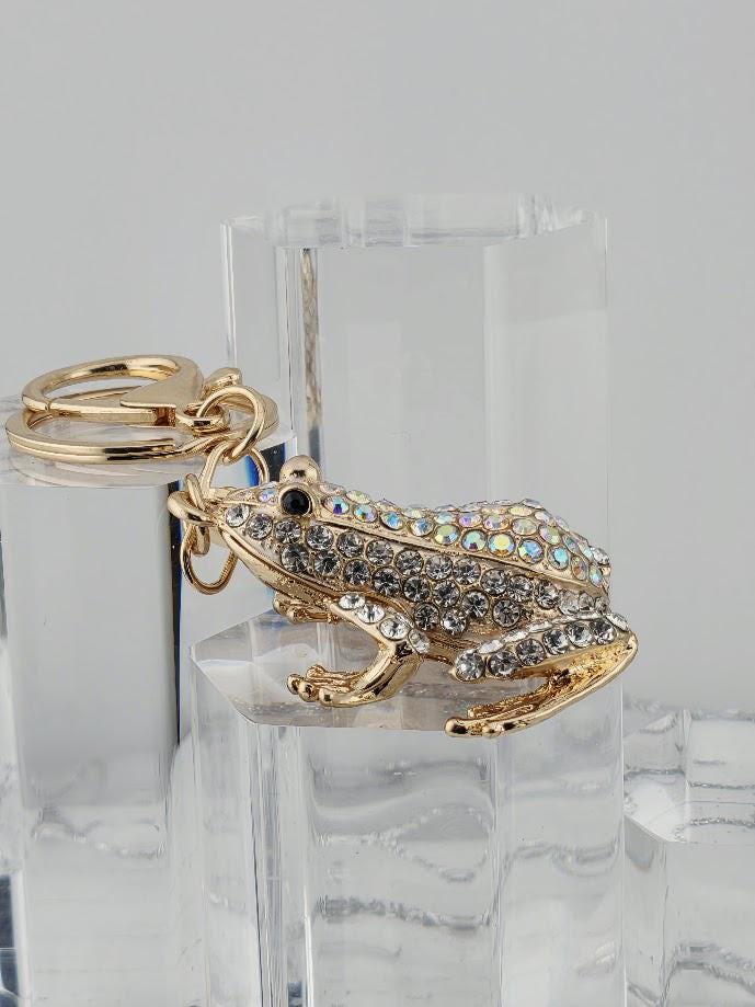A gold-colored keychain in the shape of a frog, adorned with rhinestones or crystals, is displayed in a black box.