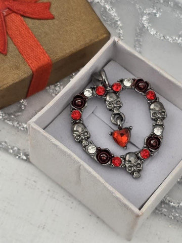 A silver necklace with red gemstones and skull-shaped charms is displayed in a white box, with a brown gift box and a red ribbon visible in the background.