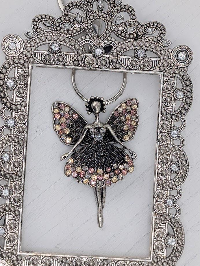 A silver-colored pendant featuring a stylized fairy figure with wings and a flowing dress, adorned with numerous small, colorful gemstones.