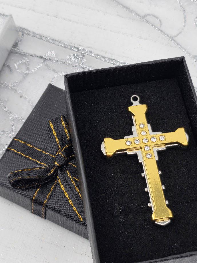 A gold cross pendant with rhinestones is displayed in a black box with a gold ribbon bow.