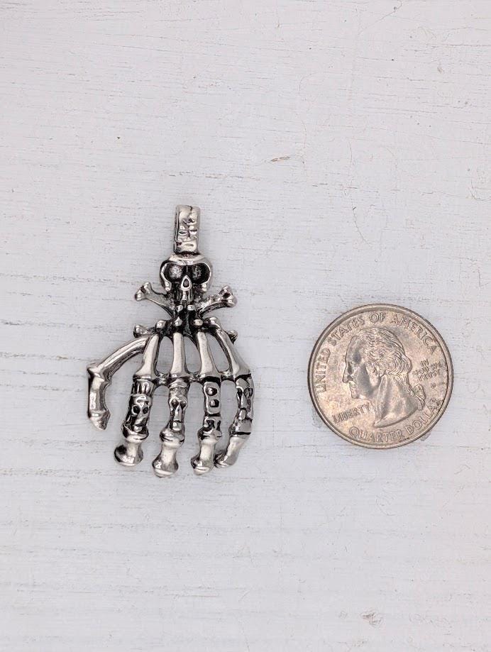 A silver-colored pendant in the shape of a hand with bones protruding from it, resting on a dark surface.
