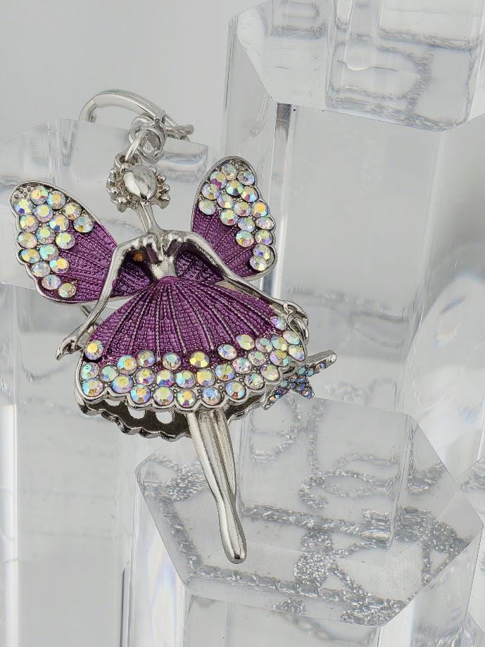 A silver-colored pendant in the shape of a fairy with wings and a purple dress, adorned with rhinestones or crystals.
