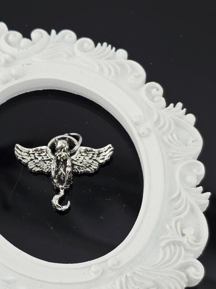 A silver-colored angel charm with wings is displayed against a black background, encased within an ornate white frame.