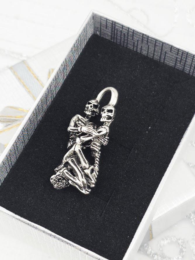 A silver-colored pendant in the shape of a skeleton embracing another skeleton, displayed in a black box with a white border.