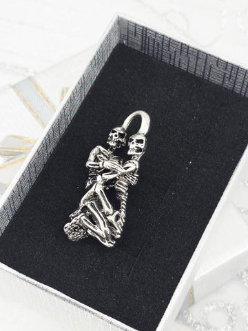 A silver-colored pendant in the shape of a skeleton embracing another skeleton, displayed in a black box with a white border.