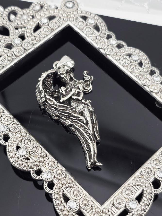 A silver-colored pendant in the shape of a winged figure is displayed against a black background, framed by an ornate, diamond-encrusted frame.
