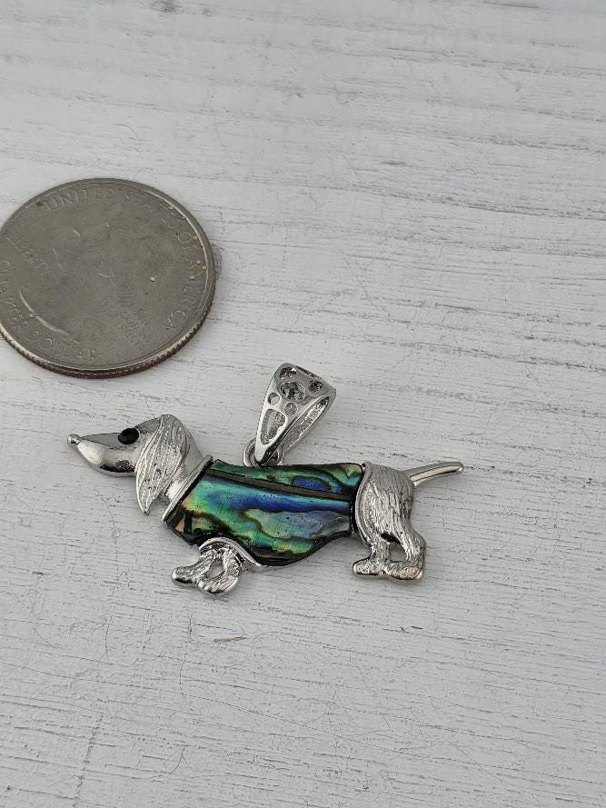 A silver pendant in the shape of a dachshund dog, with a colorful iridescent pattern, is placed next to a United States quarter on a wooden surface.