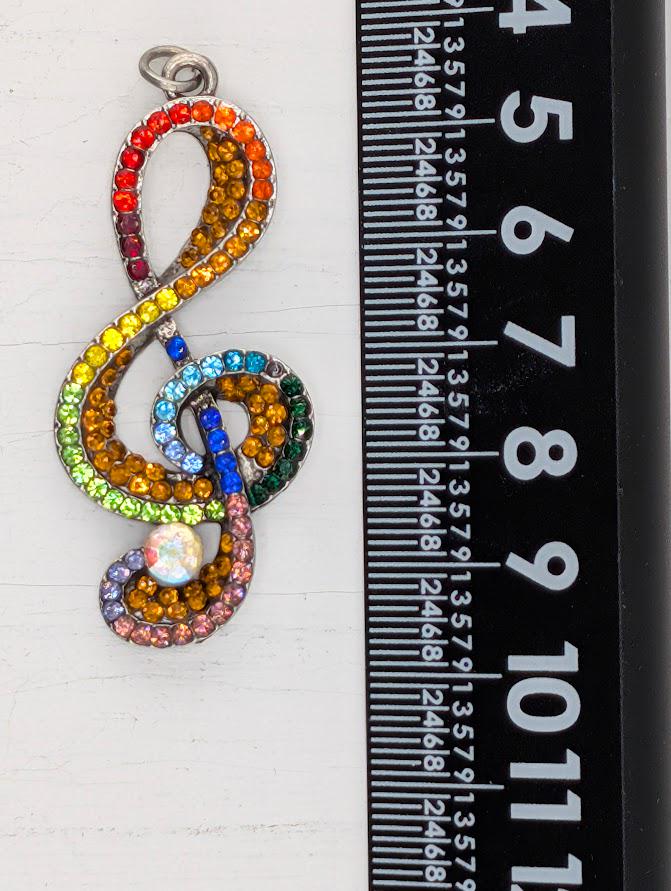 A colorful pendant with a treble clef design, adorned with multicolored gemstones, is placed next to a ruler for size comparison.