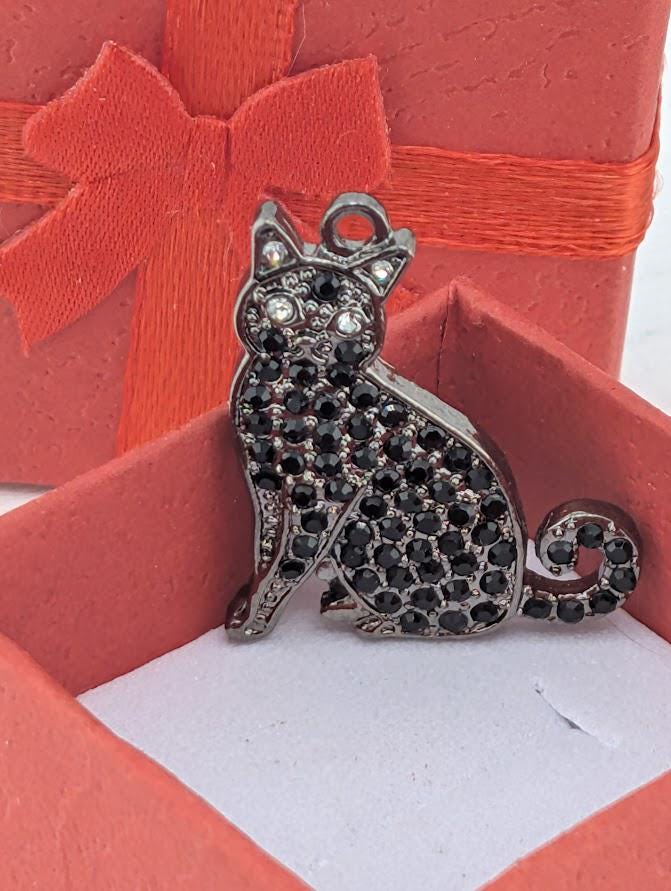 A silver-colored pendant in the shape of a cat, adorned with black gemstones, is displayed in a red gift box.