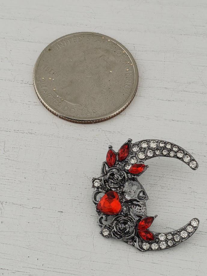 A silver brooch with a crescent moon design and red gemstones is displayed in a white box, with a brown gift box with a red ribbon visible in the background.