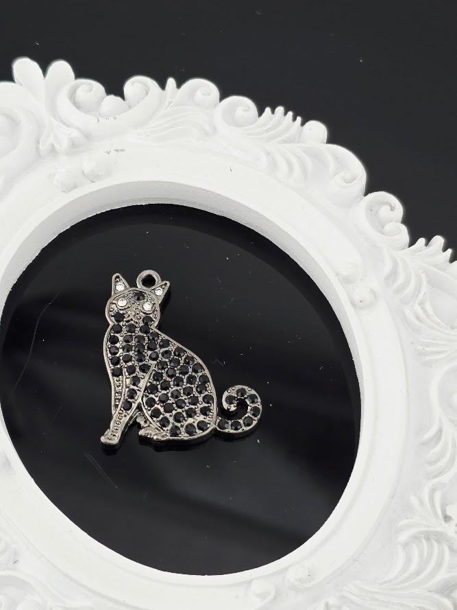 A silver cat charm with black gemstones is displayed in a white ornate frame against a black background.