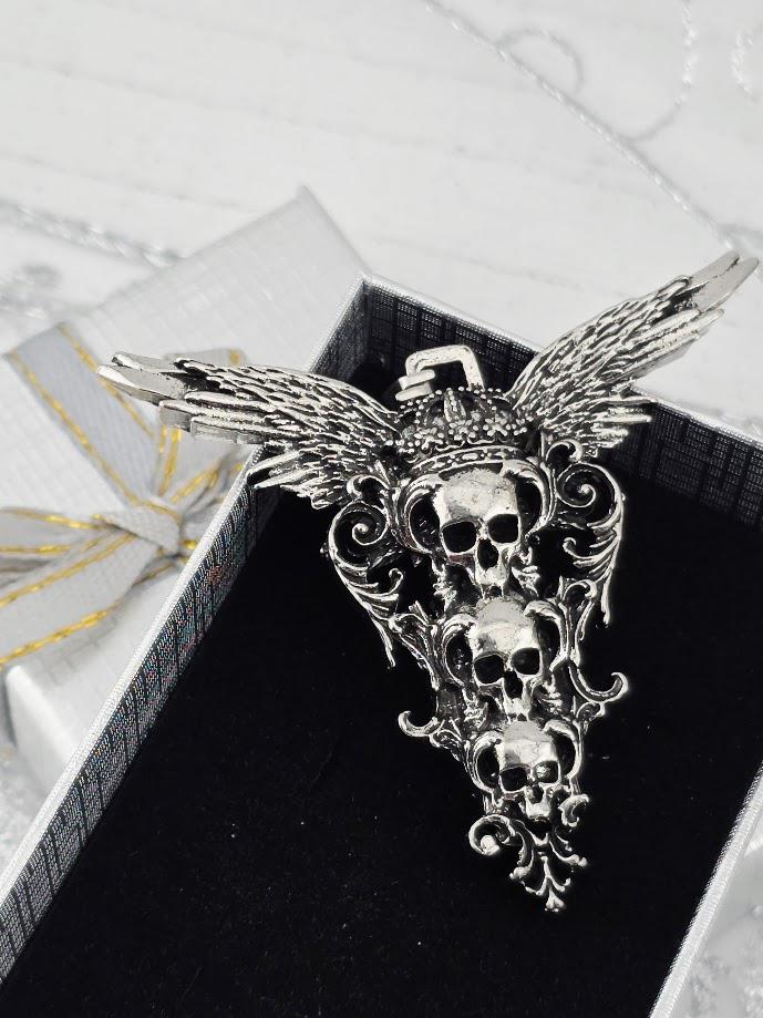A silver pendant with three skull designs and wings, displayed in a black box with a white ribbon.