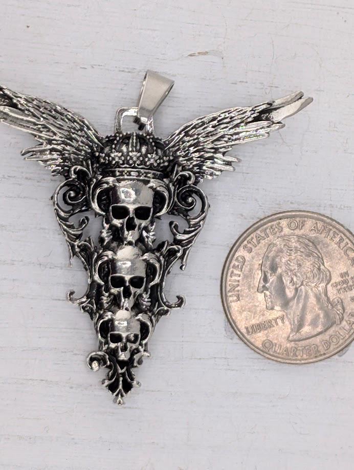 A silver pendant with three skull designs and wings, and a United States quarter coin next to it.