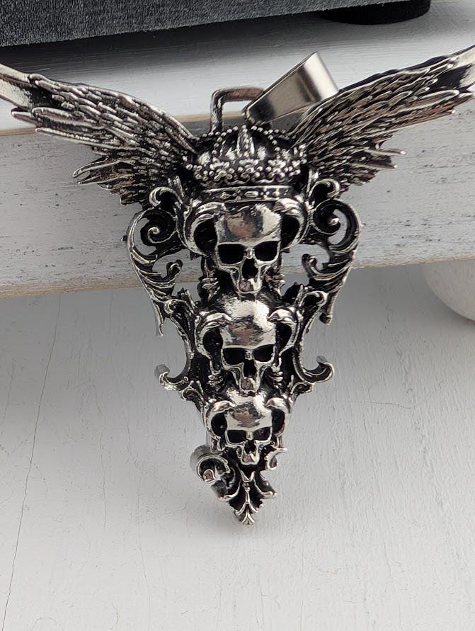 A silver pendant with a design featuring three skull faces with wings, possibly resembling a crown or a symbol of power.