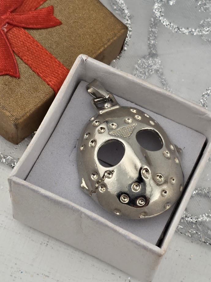 A silver pendant in the shape of a mask is displayed in a white box, with a gift box and a red ribbon visible in the background.