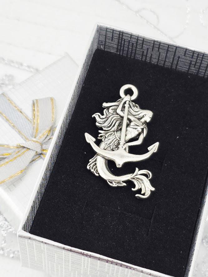 A silver mermaid-shaped pendant with an anchor is displayed in a white box with a gold ribbon bow.