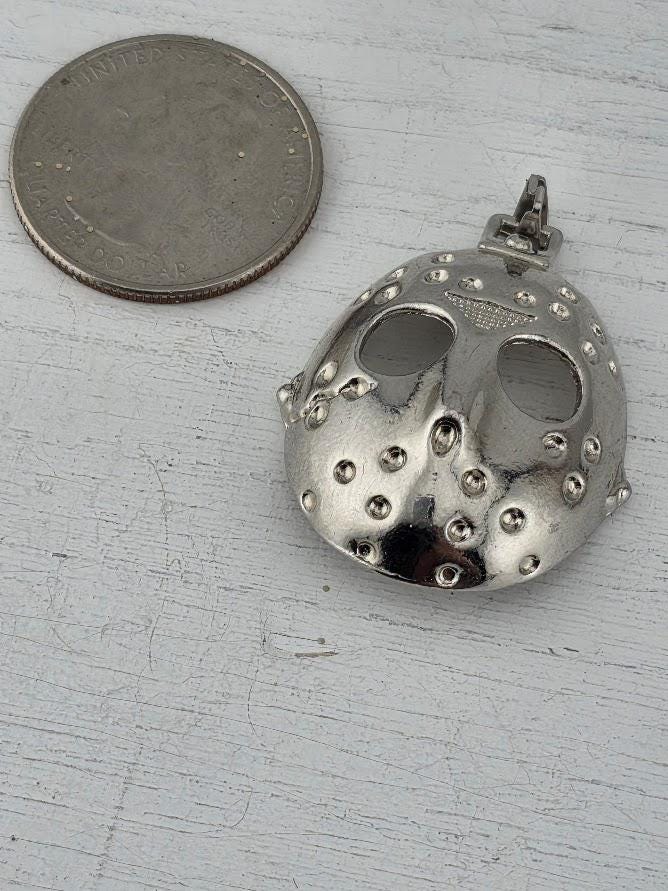 A silver pendant in the shape of a mask is displayed in a white box, with a gift box and a red ribbon visible in the background.