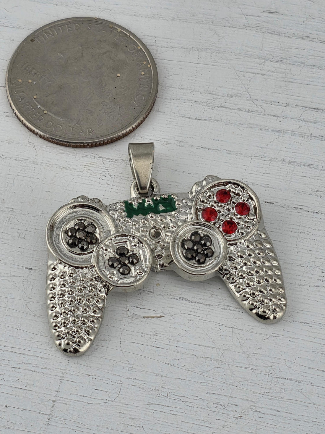 A silver and green pendant in the shape of a video game controller, with red gemstones, is displayed in a white box with a red ribbon.