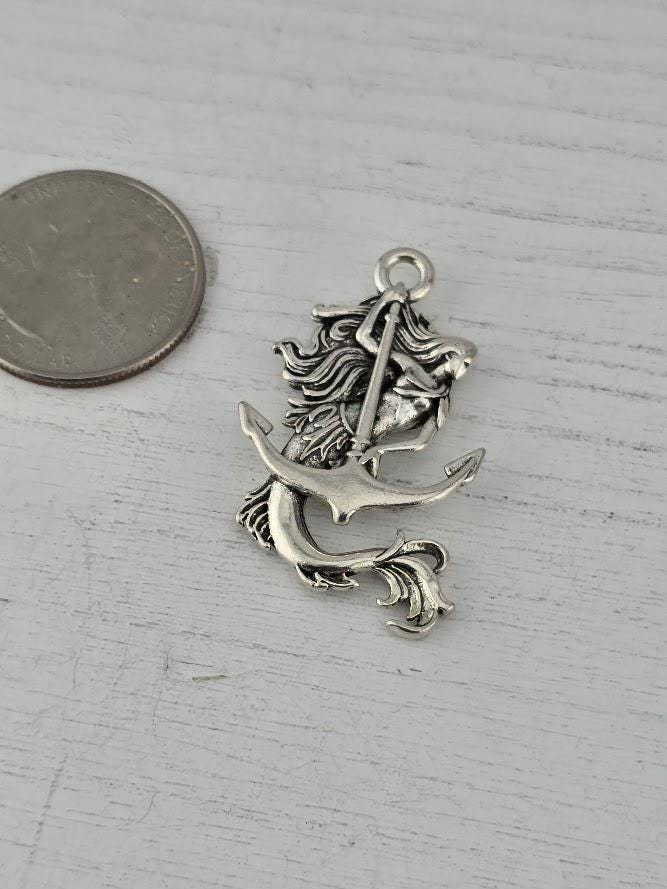 A silver-colored mermaid charm is shown, with a coin placed next to it on a wooden surface.