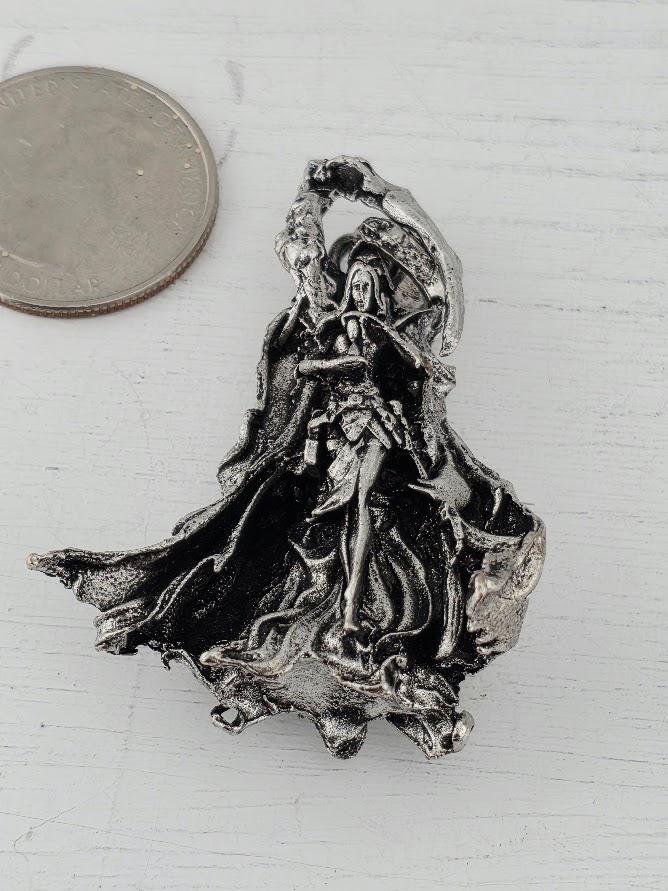 A silver-colored metal pendant in the shape of a female figure, possibly a deity or goddess, is shown next to a coin.