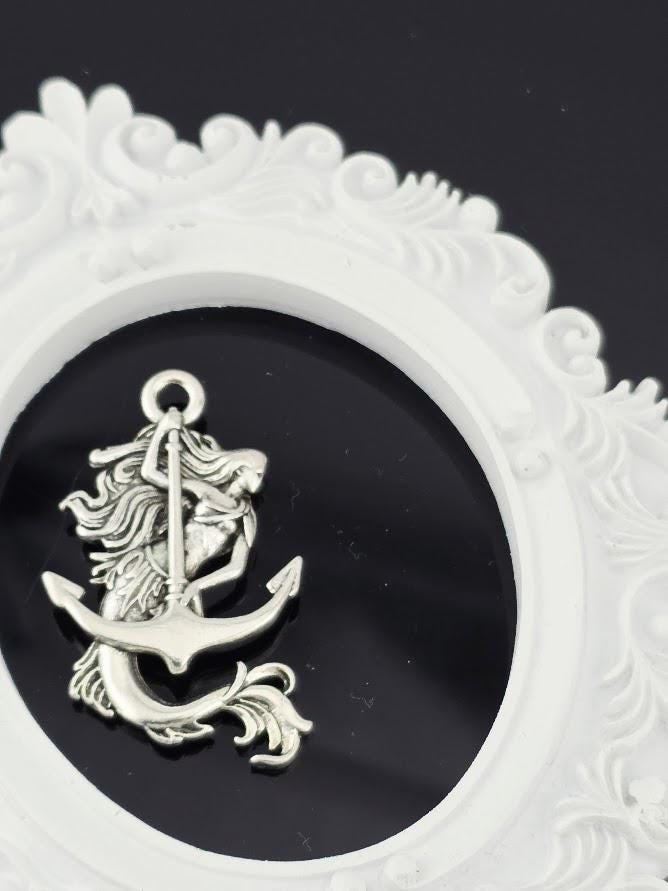 A silver mermaid charm is displayed against a dark background, encased within an ornate white frame.