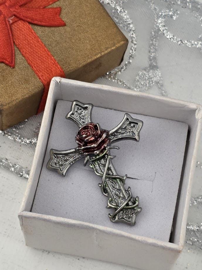 A silver cross brooch with a red rose is displayed in a white box, accompanied by a brown gift box with a red ribbon.