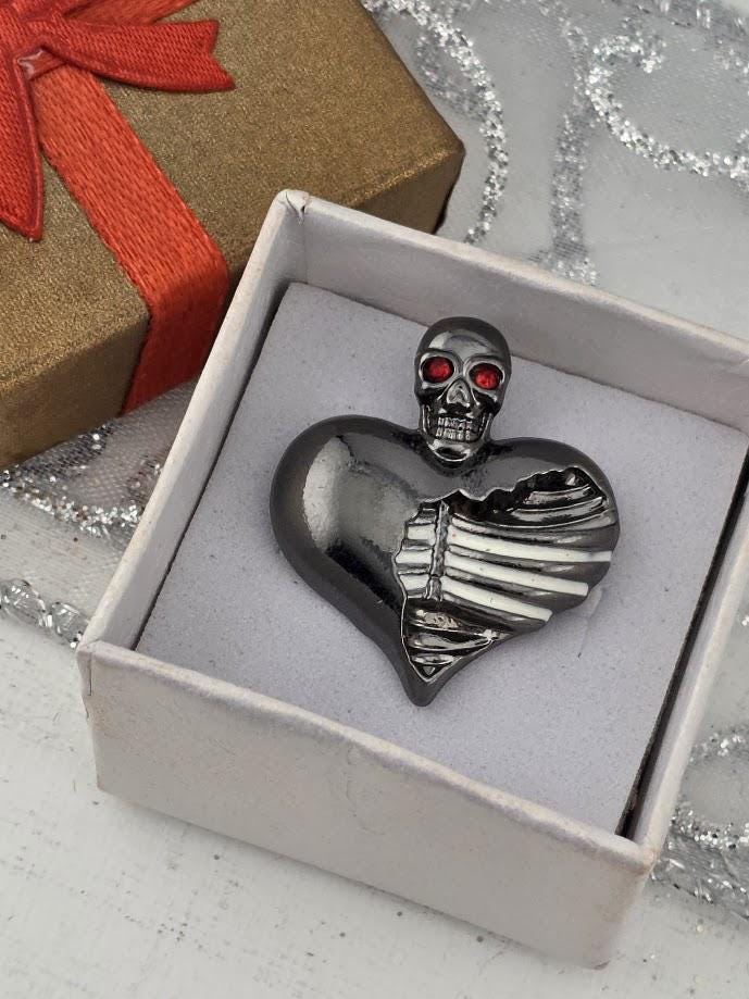 a heart-shaped brooch with a skull design, placed inside a white box.