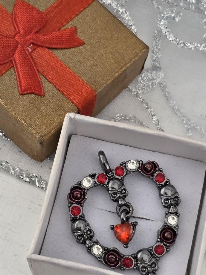 A decorative pendant in the shape of a heart, adorned with red and silver elements, is displayed in a white box with a red ribbon.