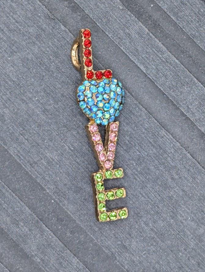A colorful pendant with the word "LOVE" made up of various colored gemstones, including blue, pink, green, and red.