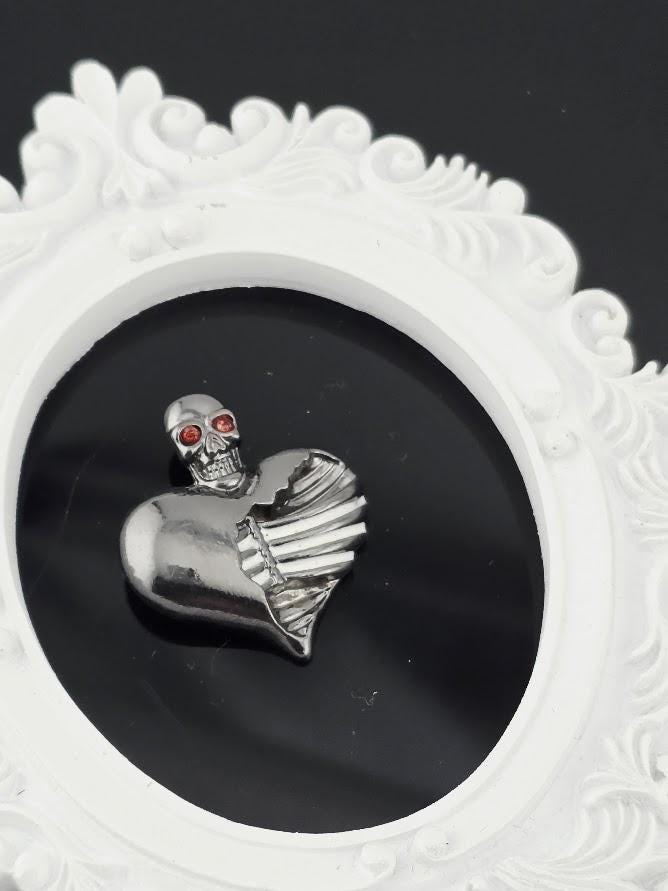A silver skull-shaped pendant with a heart-shaped cutout is displayed against a black background, encased within a white ornate frame.